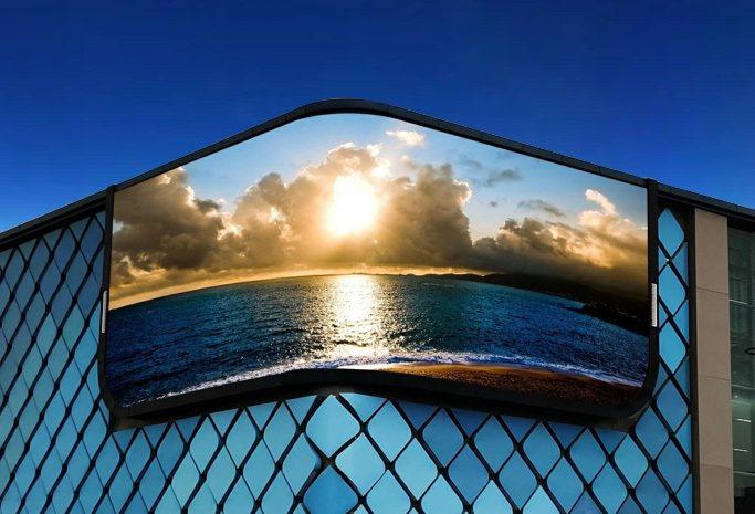 Curved outdoor LED screen set into a diamond-patterned building facade, showing a sunset sky